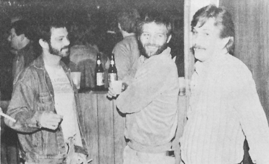 Three men stand together in a bar. Other bar patrons are in the background.