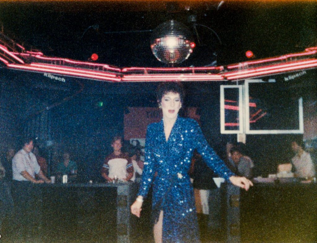 A performer in a blue sequin dress dances under a disco ball at a club.