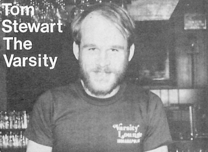 A man stands in a bar. His tshirt has a logo for the Varsity Lounge and text on the photo reads "Tom Stewart The Varsity."