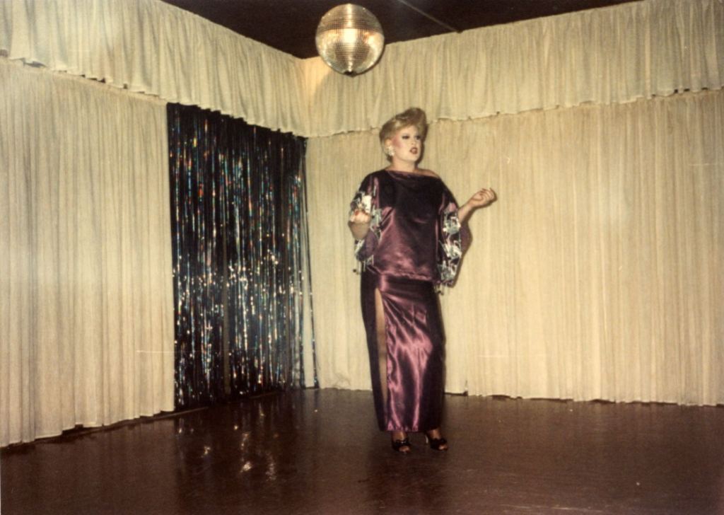A drag performer in a shiny purple dress stands on a stage.