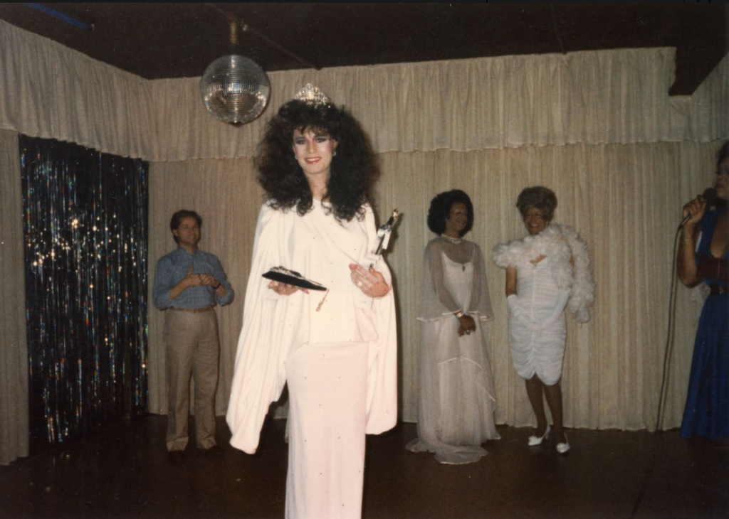 A drag performer stands at the front of a stage and holds a trophy. Other drag performers stand in line in the background.