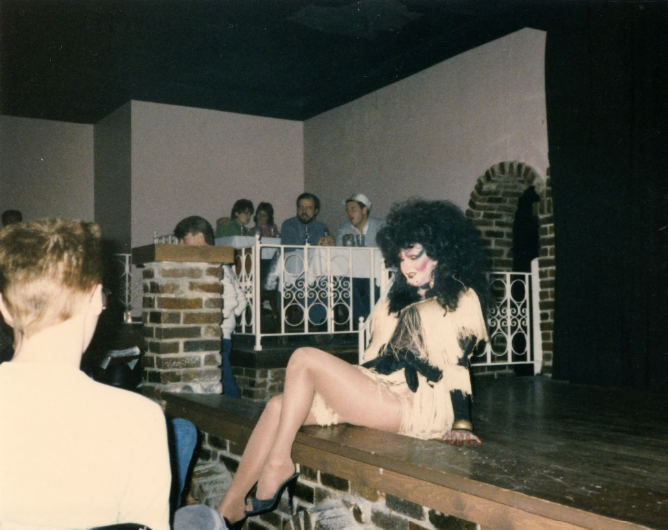 A drag performer sits on stage with audience visible in the back.
