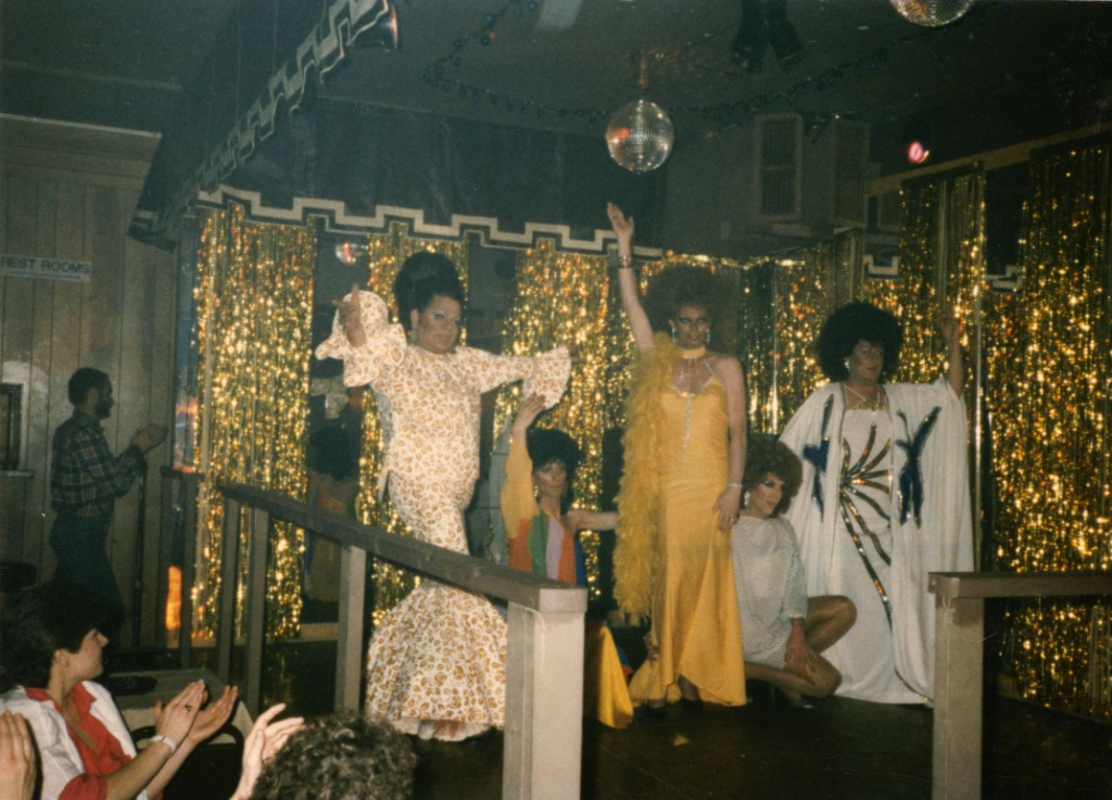 Five drag performers pose on stage. The audience watches and claps.