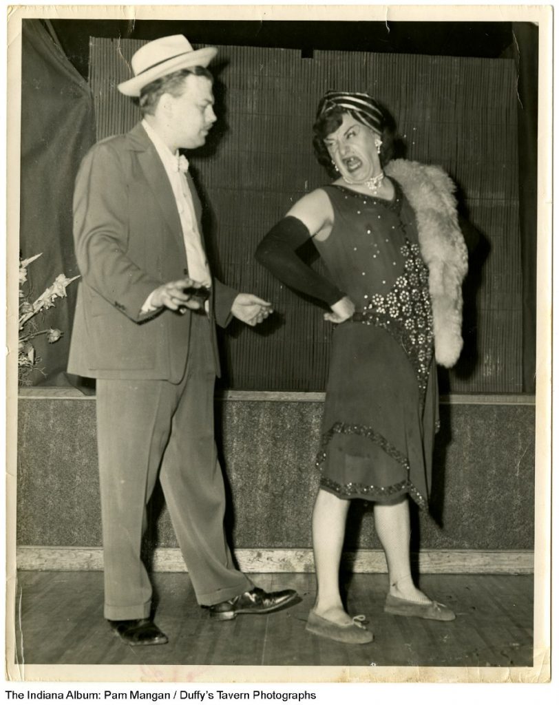 A man in a suit stands next to a drag performer on a stage.