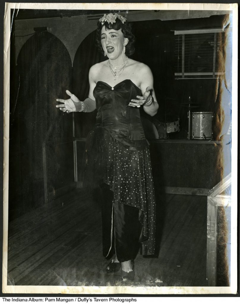 The brunette performer wears make-up, a long gown, fishnet stockings, and high heels. In the background is a drumset on a stage.