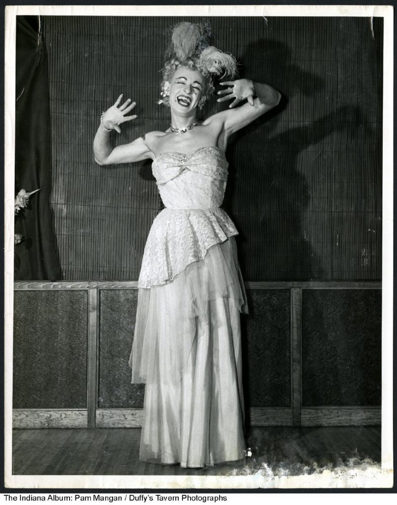 The blond performer wears a feathered hat, jewelry, and an off-the-shoulder long gown.