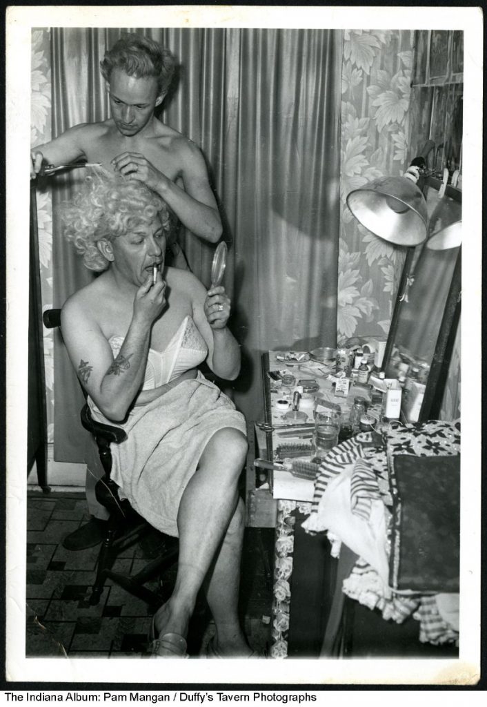 A tattooed drag performer wearing a bra applies lipstick while having his blond wig curled with a curling iron. Visible on the dressing table are brushes, scissors, an ashtray, makeup, band-aids, and other items.