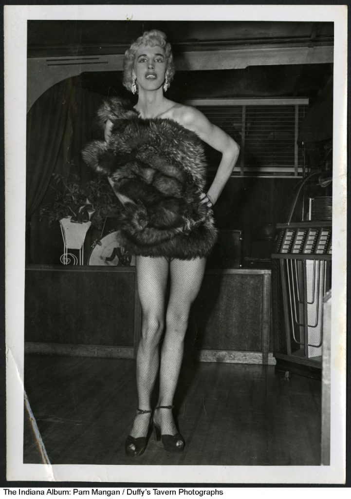 A drag performer wears makeup, a blond wig, fur over a skimpy outfit, fishnet stockings, and high-heeled strapped shoes. In the background is a stage and jukebox.