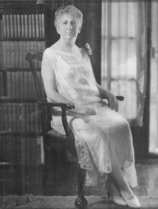 An older woman sits in a chair. She is in a room that appears to be an office or library.