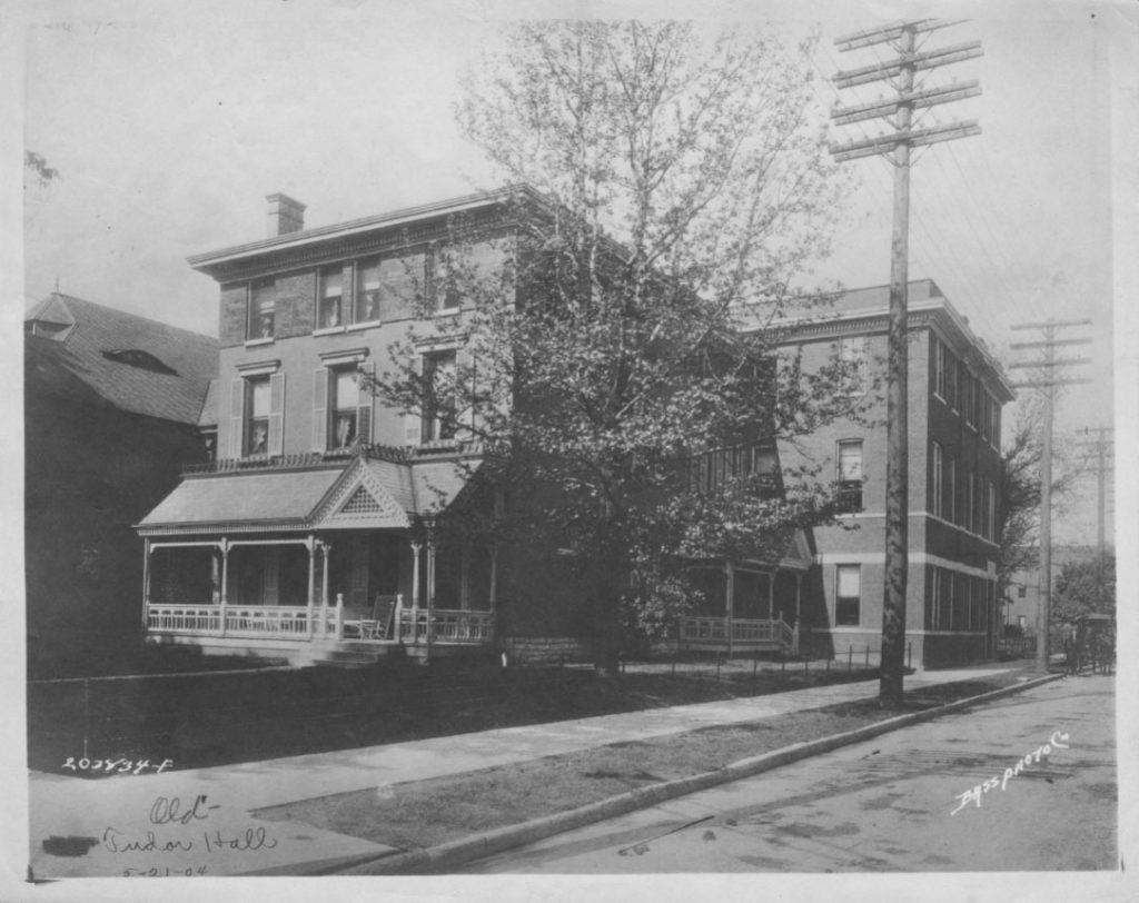 A three-story home with front porch and a side porch sits on the corner of a street. Attached to the back of the home is a square, three-story building.