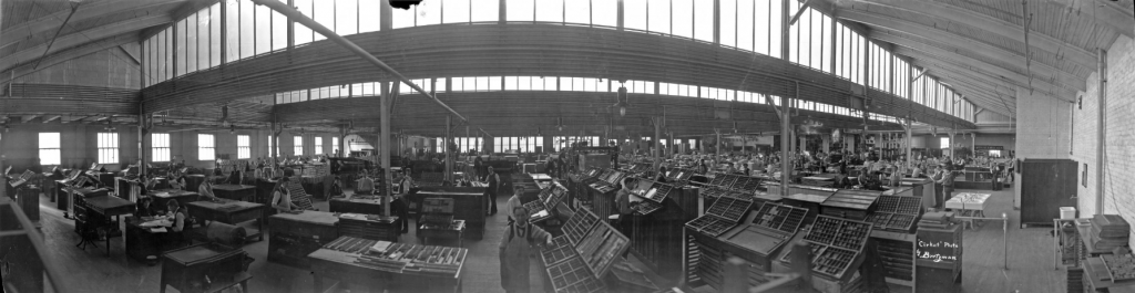 A panoramic view inside a printing press building. The large space is filled with several presses. Employees work throughout the space at individual stations.