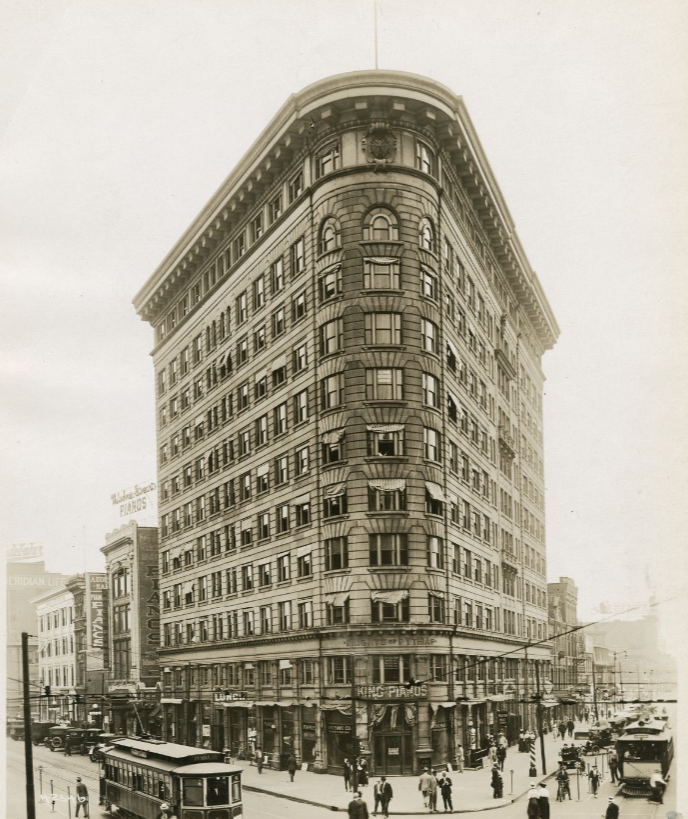 The front of an 11-story flat iron building. Street cars run on either side of the building and people walk on the side walk and in the street in front of the building.