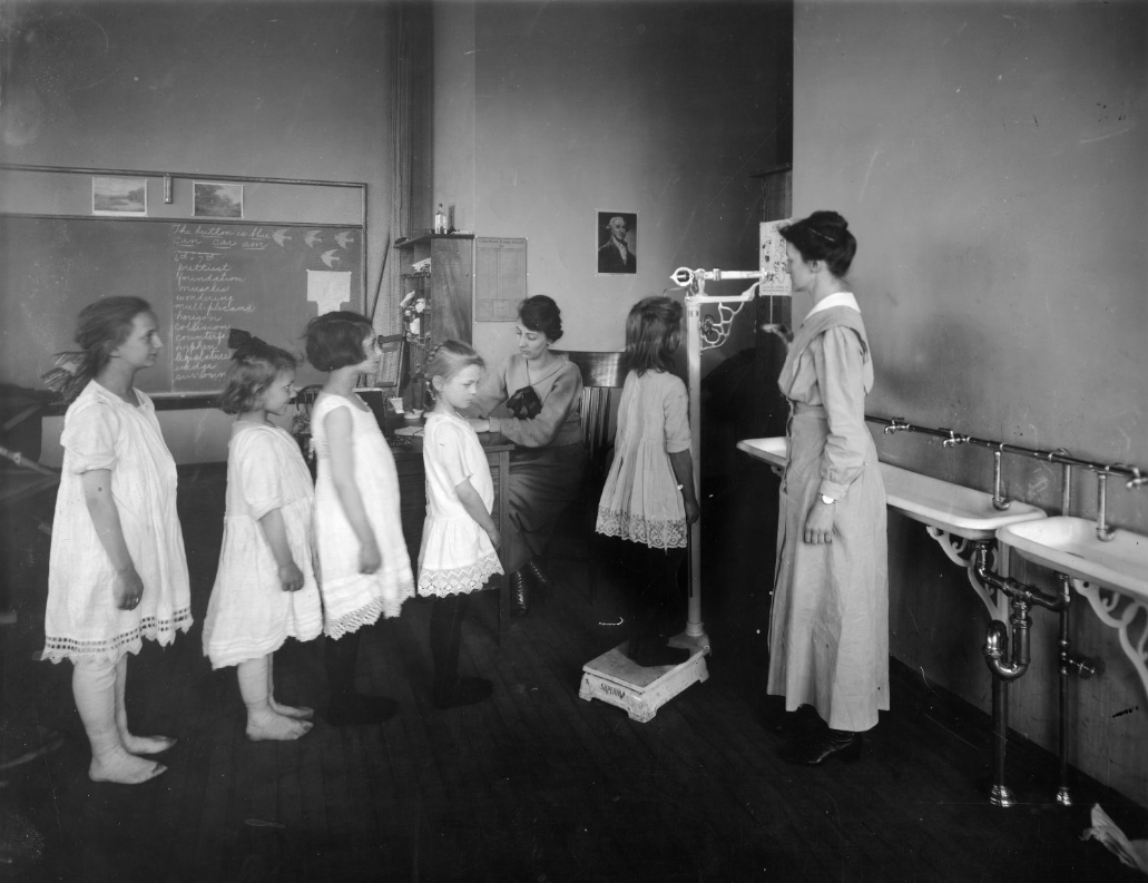 A young girl stands on a scale while one woman checks the girls weight and another woman records the weight. Four more young girls stand in line to wait their turn on the scale.