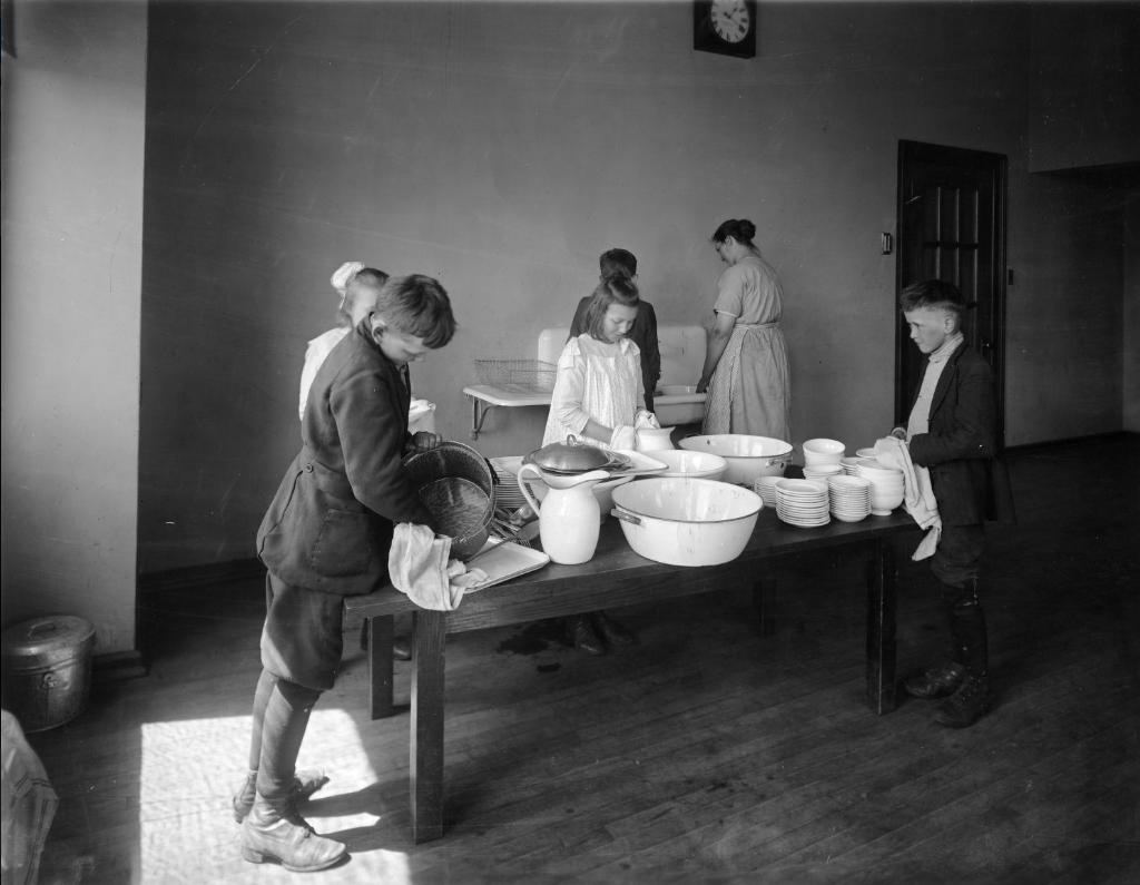 Four children work on drying dishes that are stacked on a table in the middle of a room. At the back of the room, an adult and child stand at a sink and wash dishes.