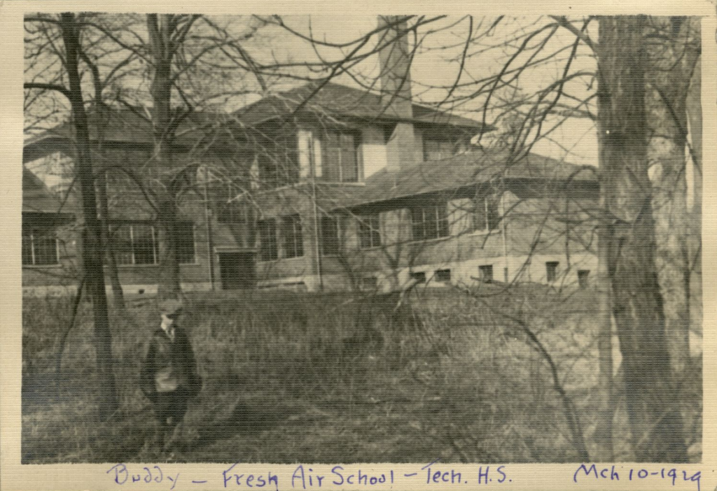 A young child stands in the tree-filled yard in front of a large house.