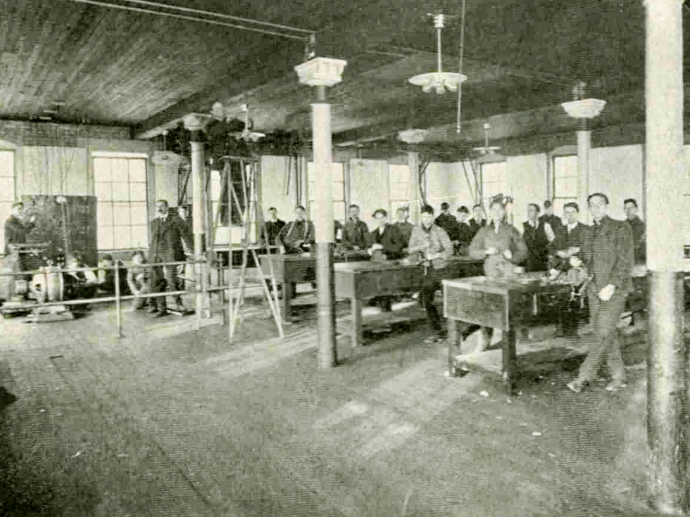 A group of male students work at four rows of tables. A large generator sits on the ground and is being worked on by several more students.