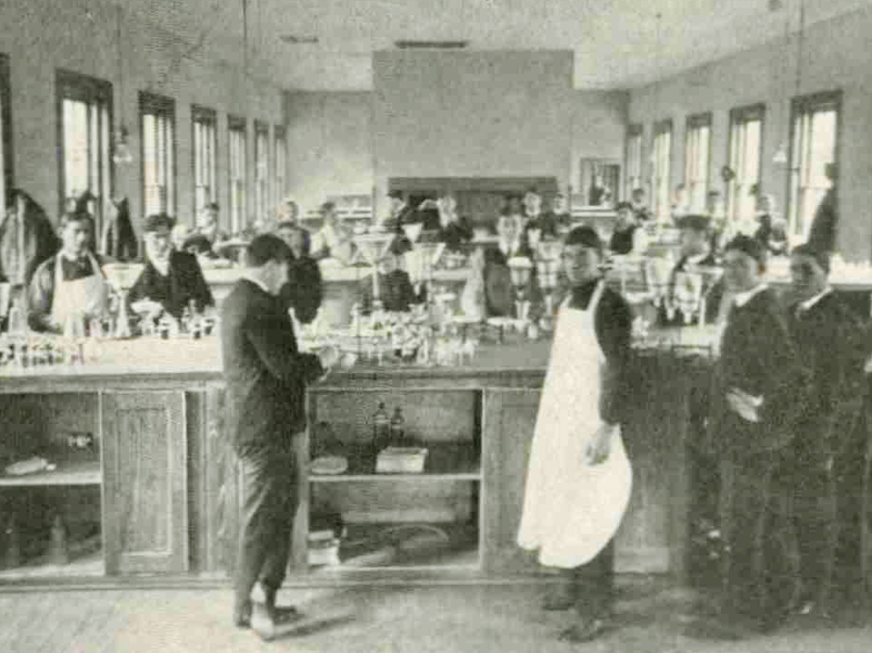 A room is filled with rows of lab tables. Students wear aprons and work with lab equipment at each of the tables.