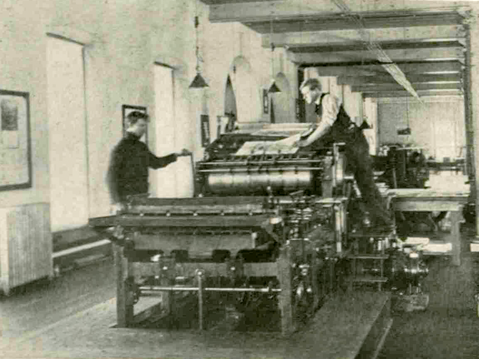 Two male students work a large printing press.