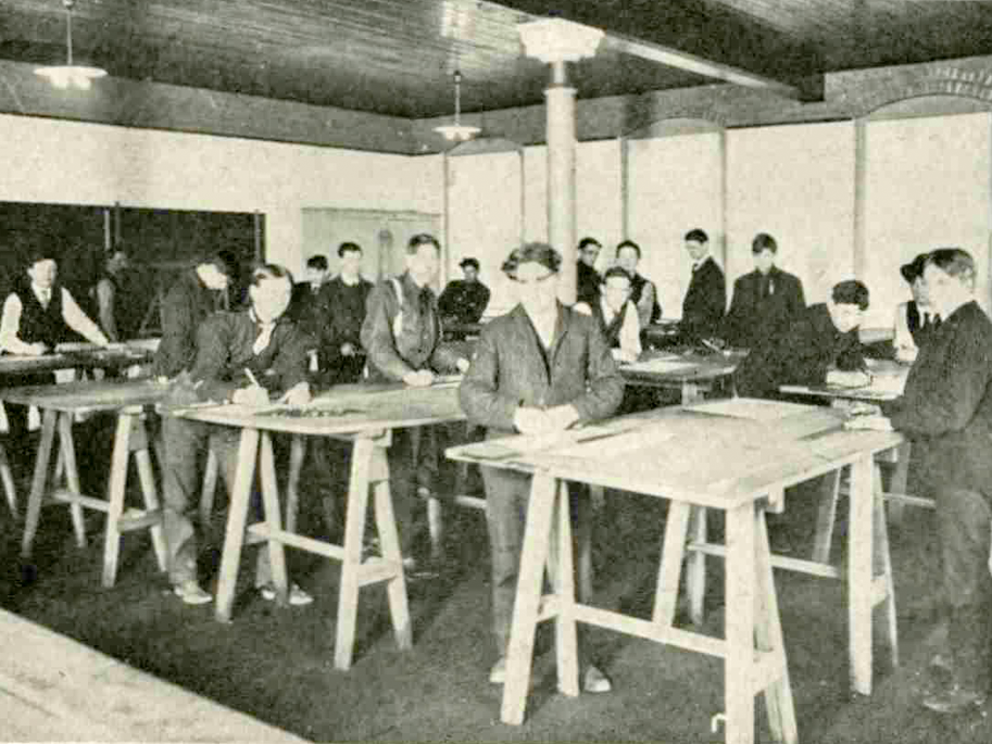 A group of male students stand at desk and work on drawings.