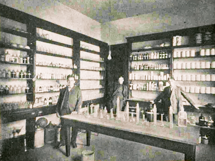 Three male students stand in a room. A table in the middle of the room as well as shelving along the walls are all filled with bottles, vials, and containers.
