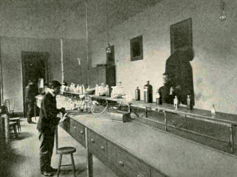 A long lab table sits in the middle of a room. Two students work on one side of the table and one more student works on the other side of the table. Various bottles are scattered along the middle of the table.