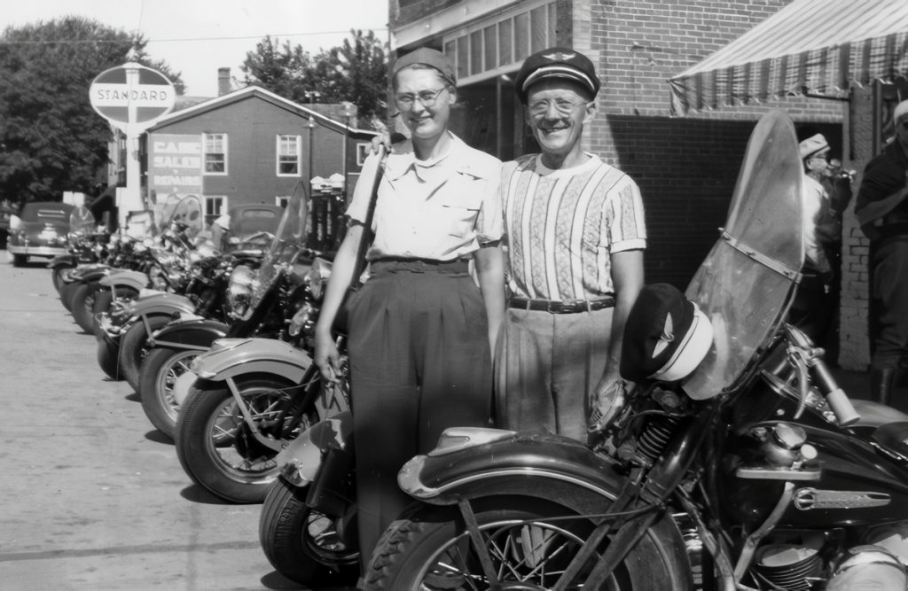 A man and a woman stand amongst a row of motorcycles lined up outside of a building.