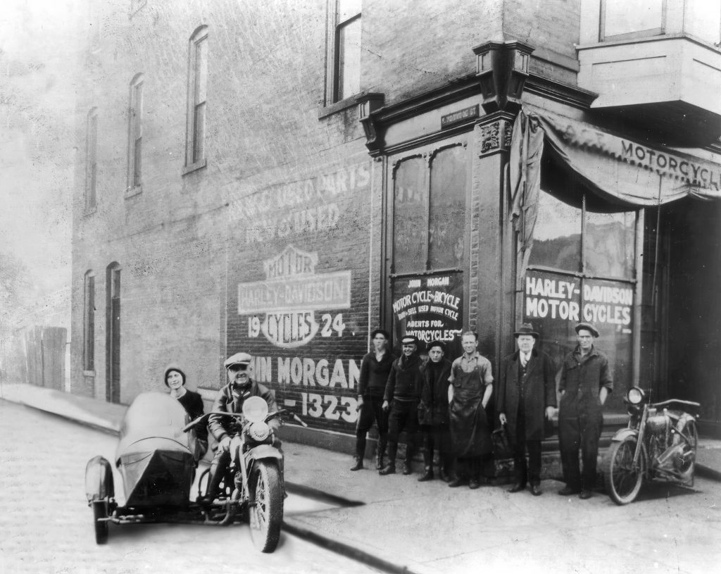 A group of people stand together outside of a motorcycle shop. Parked outside of the shop is a motorcycle with an attached sidecar. A man sits on the motorcycle and a woman sits in the sidecar.