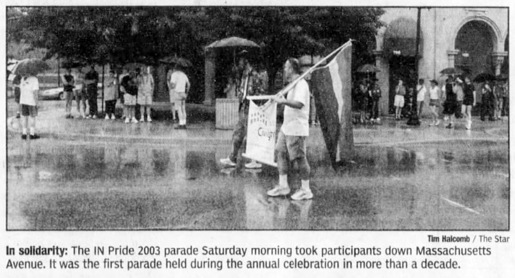 Two parage participants hold a sign while walking. One also holds a pride flag. A small group of people stand on the sidewalk and watch the parade. Several people are holding umbrellas.