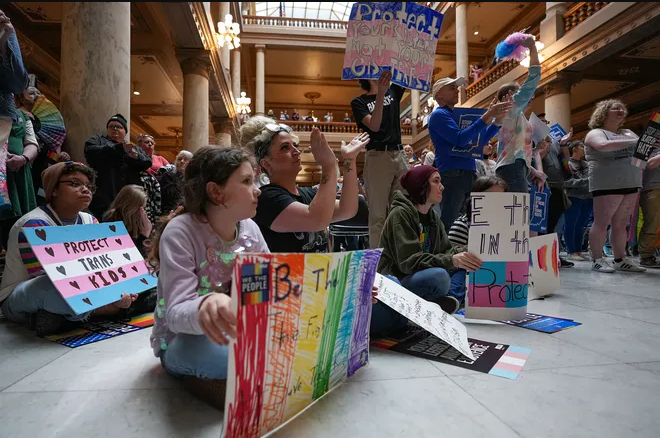 A group of adults and children sit on the floor of a room in the statehouse. Many of the people are holding signs advocating for trans children.
