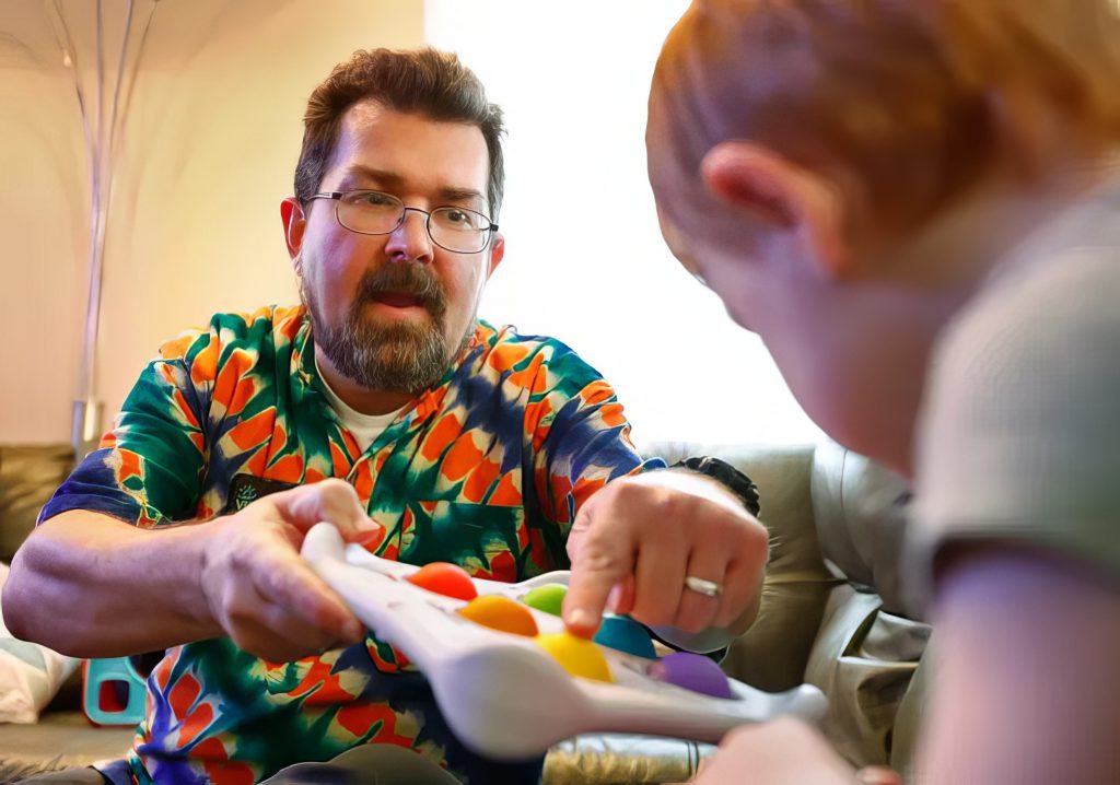A man holds out a toy to a toddler. He points to a specific spot on the toy for the toddler to focus on.