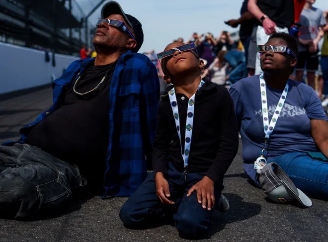 Two adults and a child sit on the track pavement and look up to the sky. All are wearing protective eyewear for the impending solar eclipse.