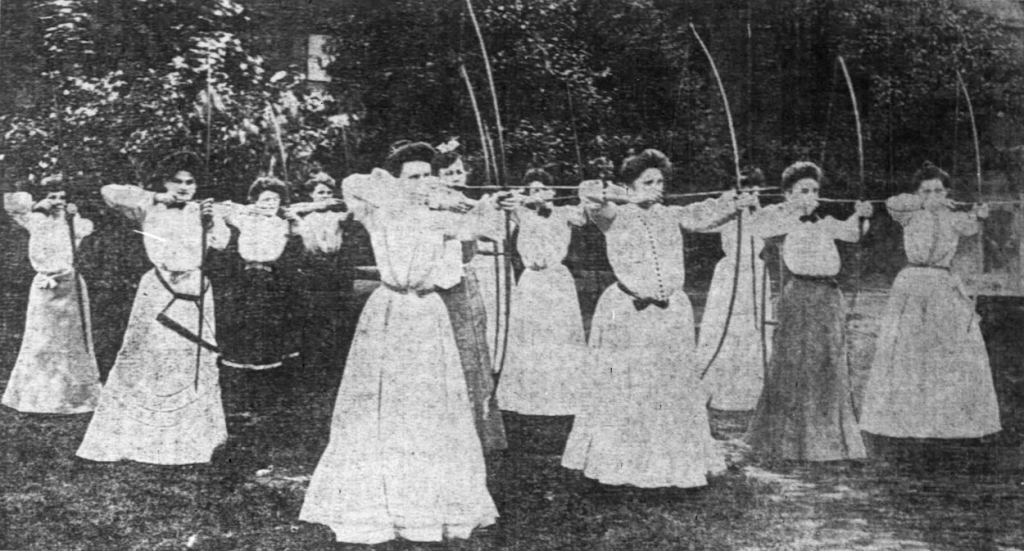 A group of young women stand in a yard. All are holding up notched bows, ready to shoot their arrows.
