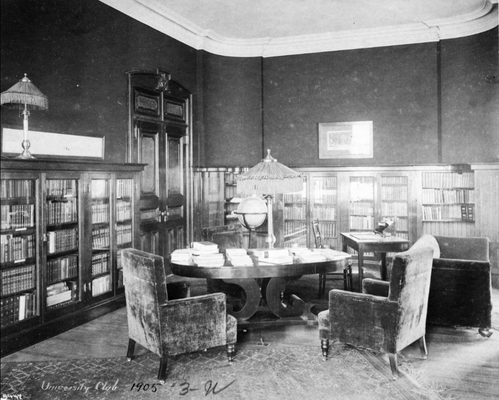 A library or study room with bookshelves lining the walls. A large table in the middle of the room is covered in stacks of books.