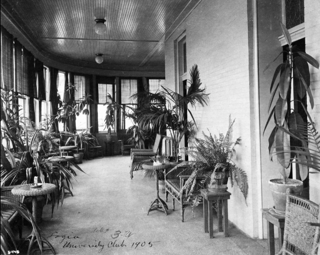 A sunroom with wicker tables and chairs lining the walls. Potted plants also line the walls.