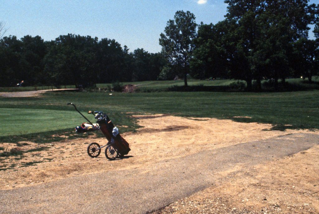 A golf bag sits in the middle of a sandy area. In the background is a grassy area and trees.