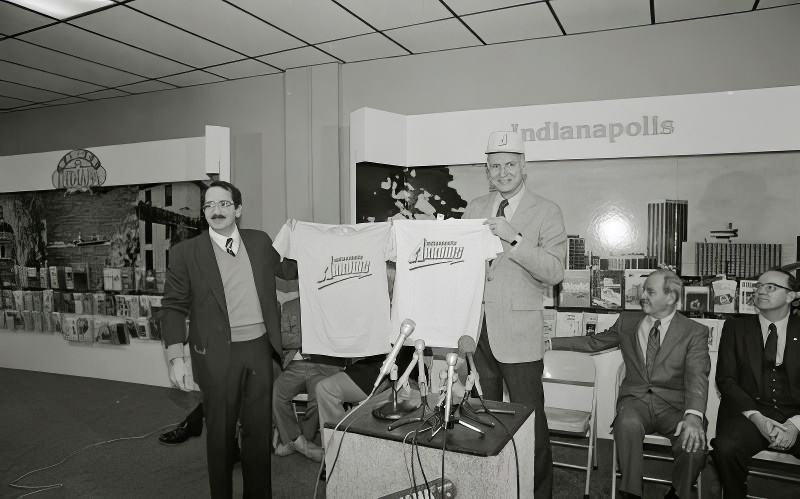 Two men hold up tshirts with the Indianapolis Arrows logo on the fronts. One man also wears a hat with the Arrows logo on the front.