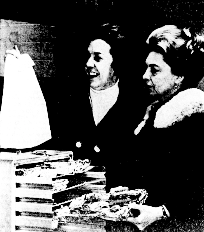 Two women stand next to each other. One woman holds up a bag and the other slides a tray of food in a box.