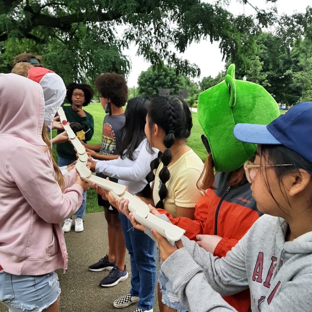 A group of children work together in a team building exercise that involves holding together individual pieces of pipe to transport a ball from one end to the other.