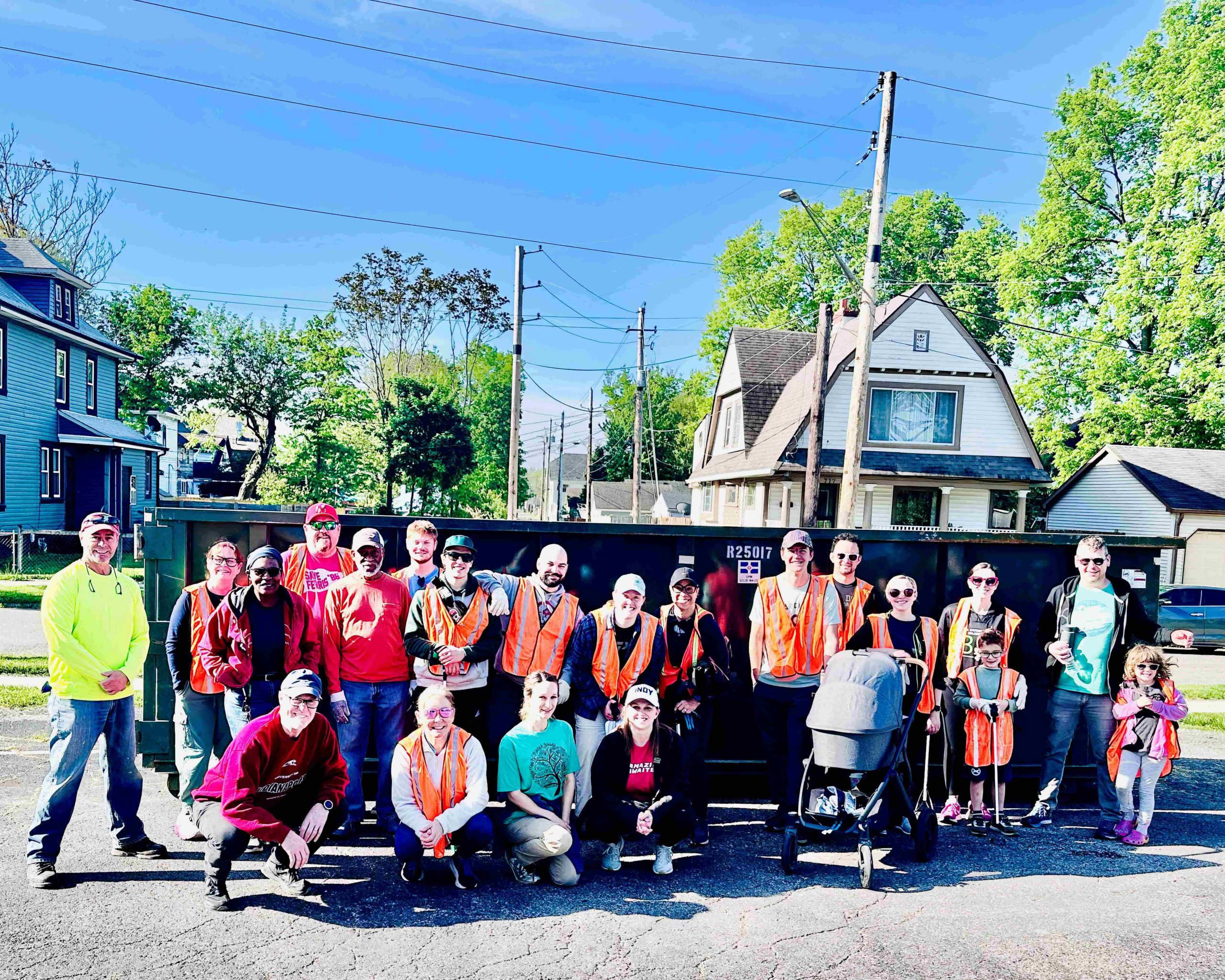 A group of people are gathered together for a photo. The majority of the people are wearing orange vests. There is a large dumpster in the background.