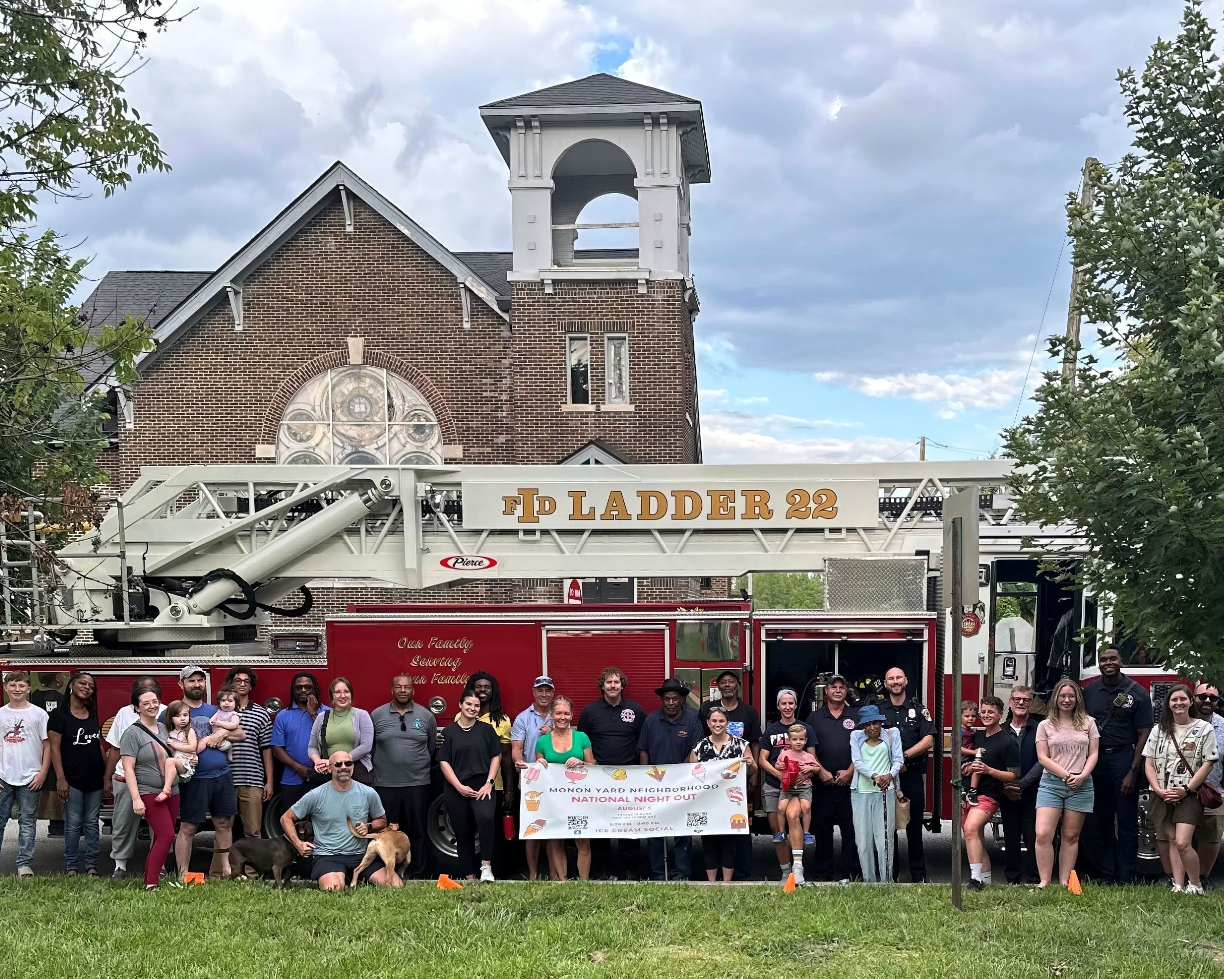 A group of people gather in front of a fire truck. The people in the middle hold up a large sign that reads "Monon Yard Neighborhood National Night Out." A church is in the background, behind the fire truck.