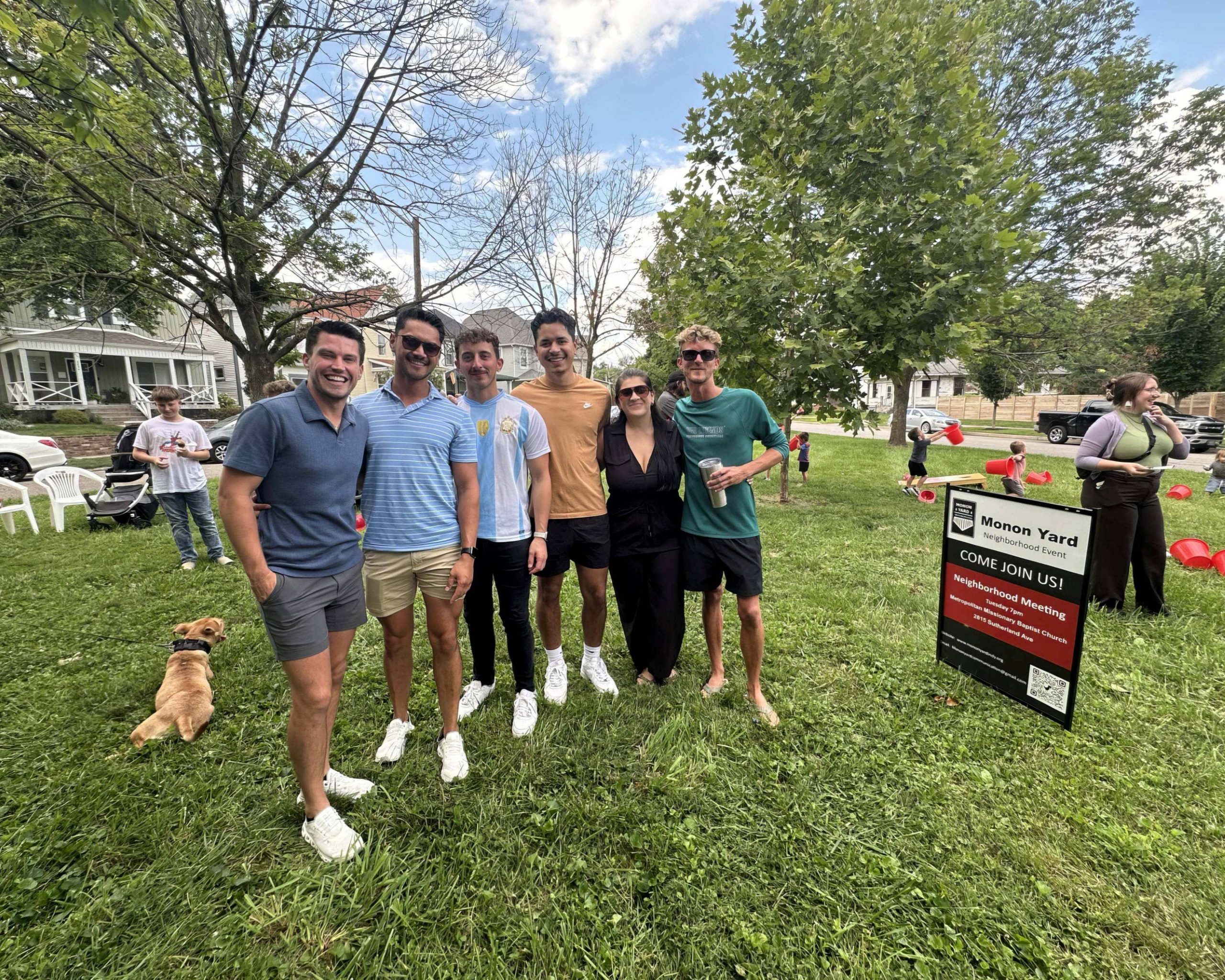 A group of people are gathered on a lawn. Some people are posing for a photo, while children play in the background. A yard sign reads "Monon Yard Neighborhood Event."