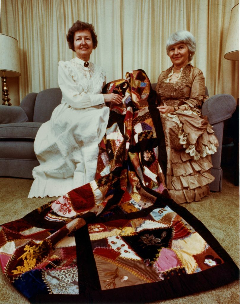 Two women dressed in early 1900s-style dresses sit together and show a large quilt.