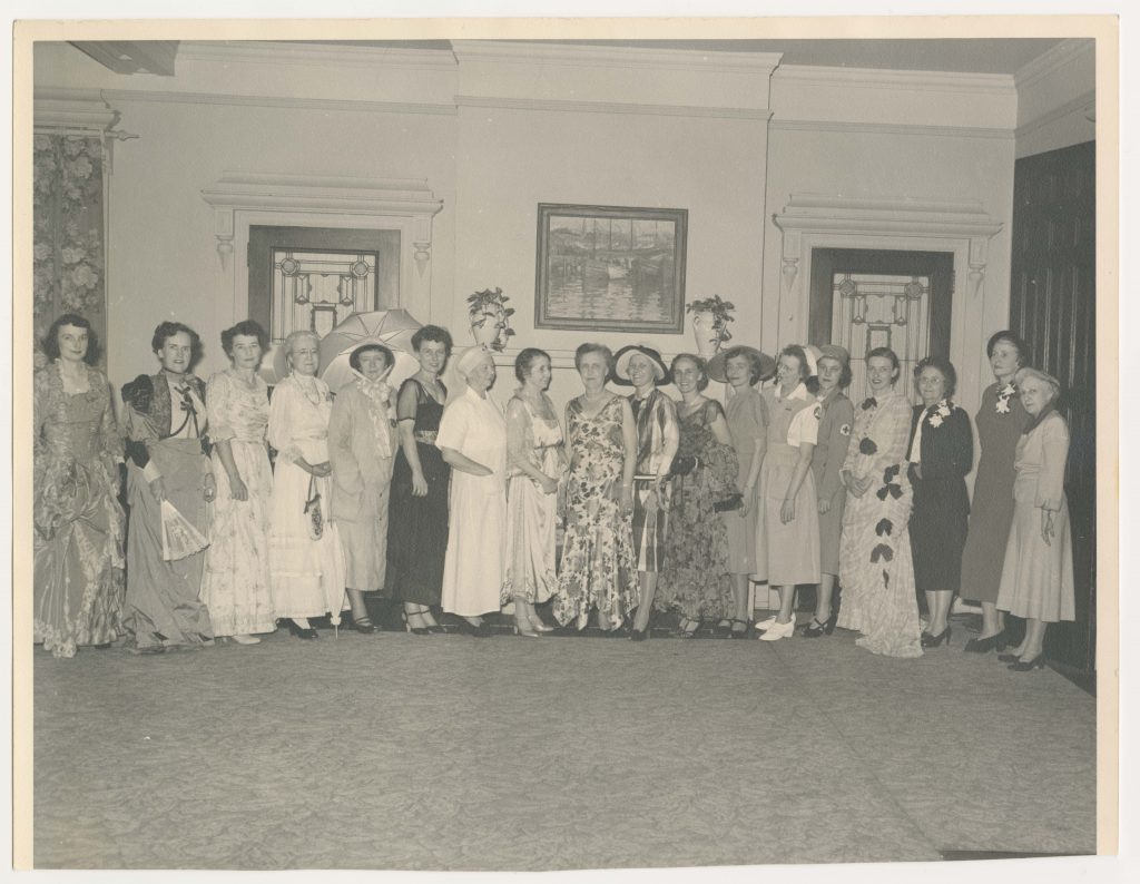 Eighteen women dressed in costumes representing different eras stand together in a line.