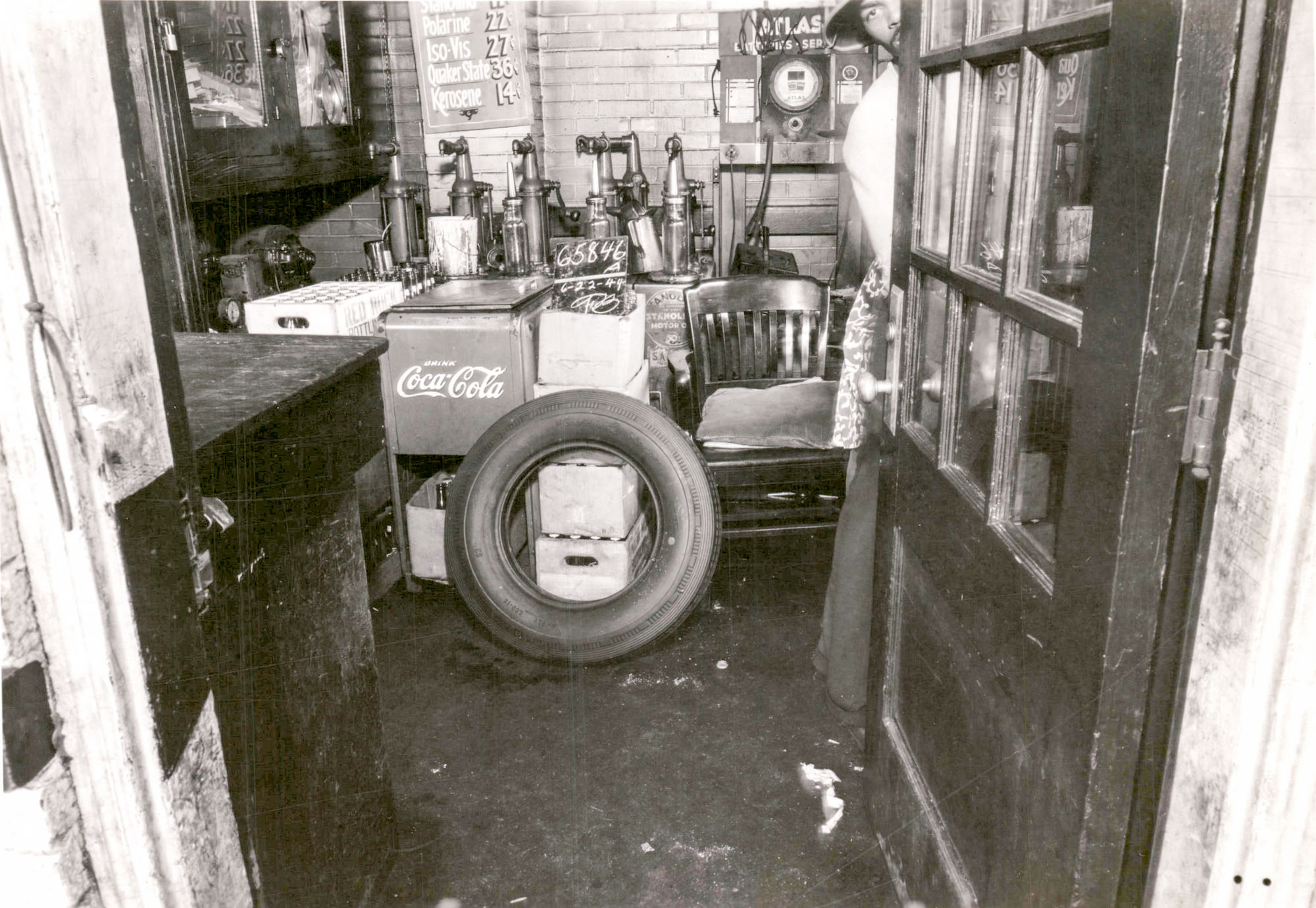 A view through the entrance to a gas station. A man is holding the door open. Cases of drinks are stacked up an a tire is leaning against them.