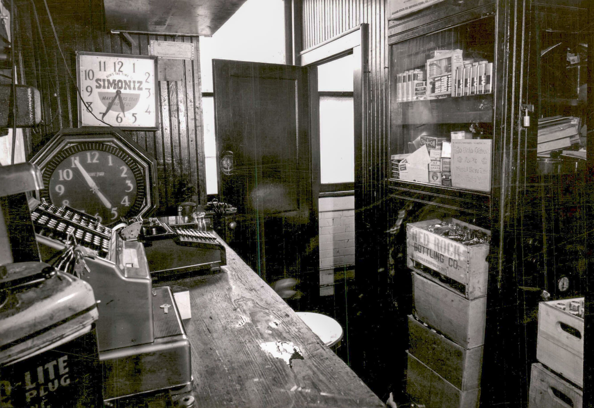 A view behind the counter of a gas station. A cash register sits on the counter, there are several clocks on the wall, and the shelves behind the counter are filled with various products.