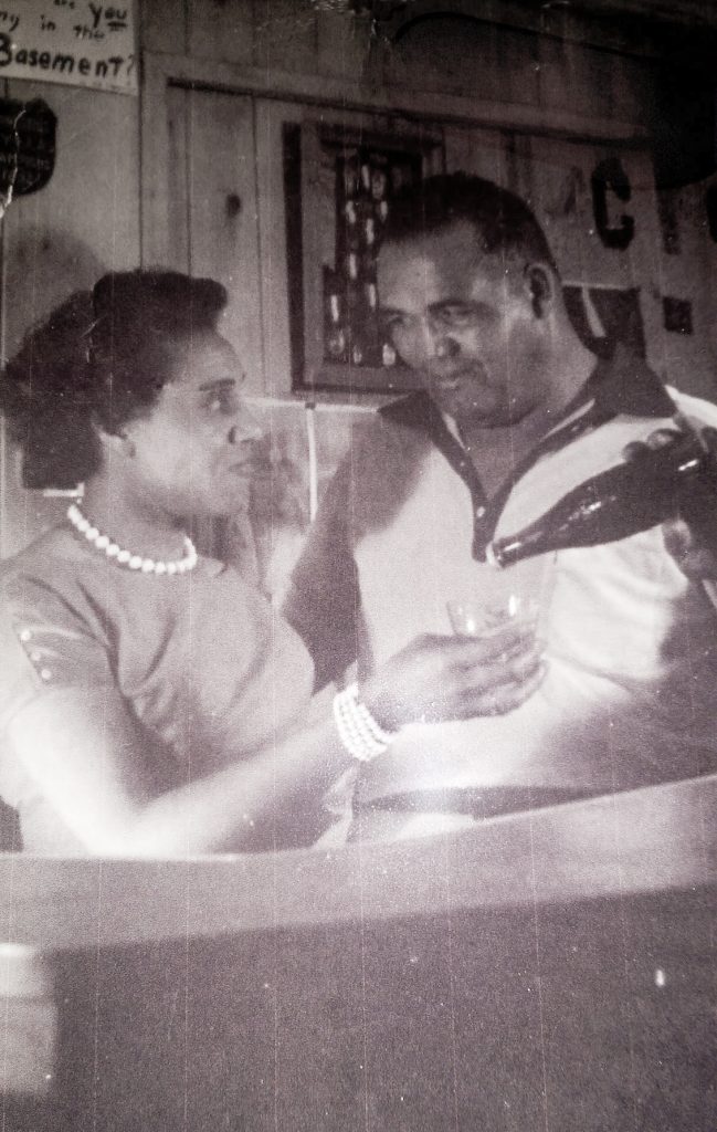 A man and a woman look into each others eyes lovingly. The woman is holding up a glass and the man is pouring her a drink.