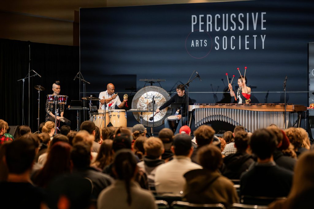 Four musicians play percussion instruments on a stage. The audience is in the foreground, watching the performance.
