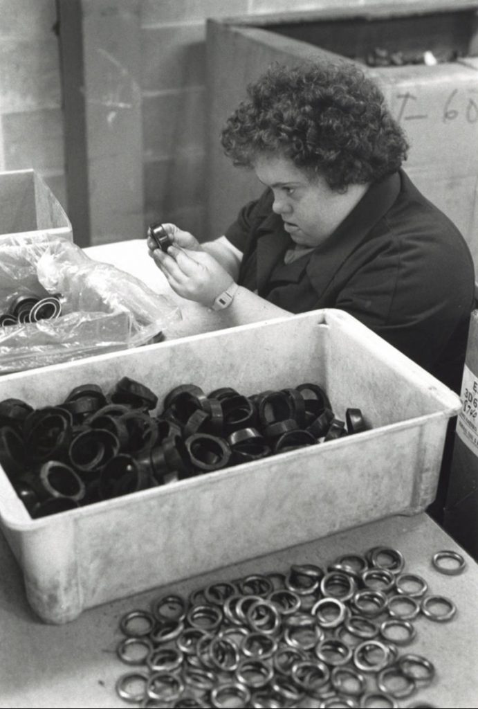 A woman with down syndrome sits at a table and puts together parts from large bins in front of her.