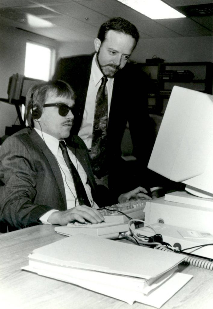 A blind man uses a computer with an assistive device. Another man leans over him.