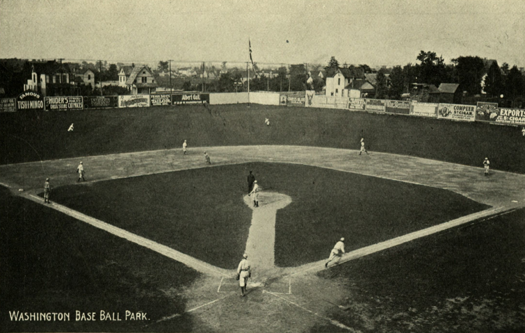 A historic baseball game at Washington Base Ball Park, viewed from behind home plate, with players on the field and advertisement signs along the outfield fence.